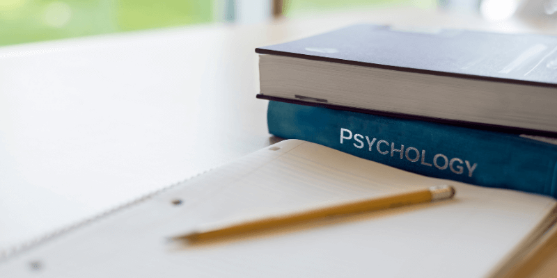 A pen and paper on a desk with a pile of psychology books.